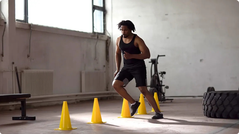 A man in a gym performs agility drills, swiftly moving between yellow cones. He's wearing a black tank top and shorts, with large tires and a stationary bike in the background.