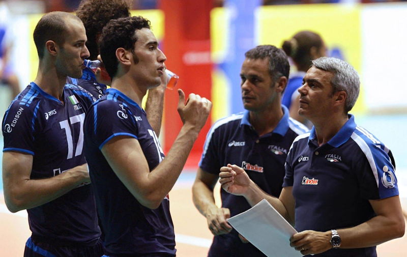 A volleyball team in blue jerseys is having a discussion with their coach, who is holding a paper. One player is drinking from a bottle, and others are listening attentively.