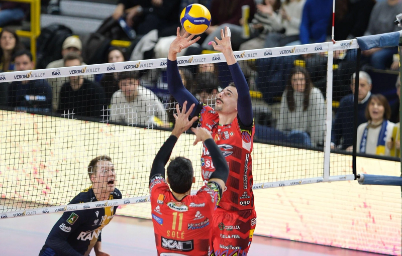 A volleyball player in a red jersey jumps to set the ball above the net, while another player in a blue jersey watches. The crowd is visible in the background.