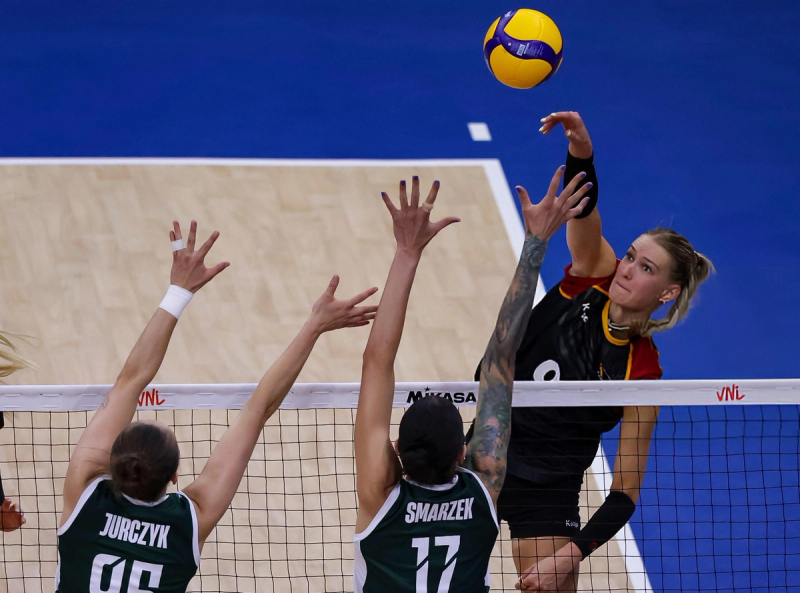 A volleyball player in a black and yellow uniform is spiking the ball over the net, while two opposing players in green jerseys attempt to block. The court is blue and the focus is on the intense action at the net.