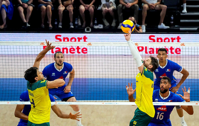 Men's volleyball match between players in yellow and green jerseys and blue jerseys. One player in a yellow jersey is reaching back to hit the ball over the net. Spectators can be seen in the background.