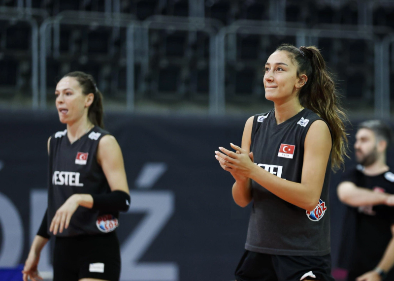 Two female volleyball players in black jerseys practice on a court. One claps while the other focuses. Turkish flags are visible on their uniforms.