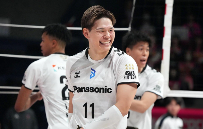 A volleyball player wearing a white "Panasonic" jersey with the number 11 is smiling on the court. The jersey sleeve also displays "Osaka Bluteon" and stars. Other players are visible in the background.