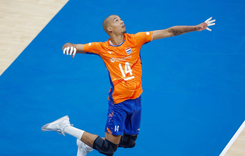 A volleyball player in an orange jersey with the number 14 is in mid-air, preparing to serve or spike the ball on a blue court.