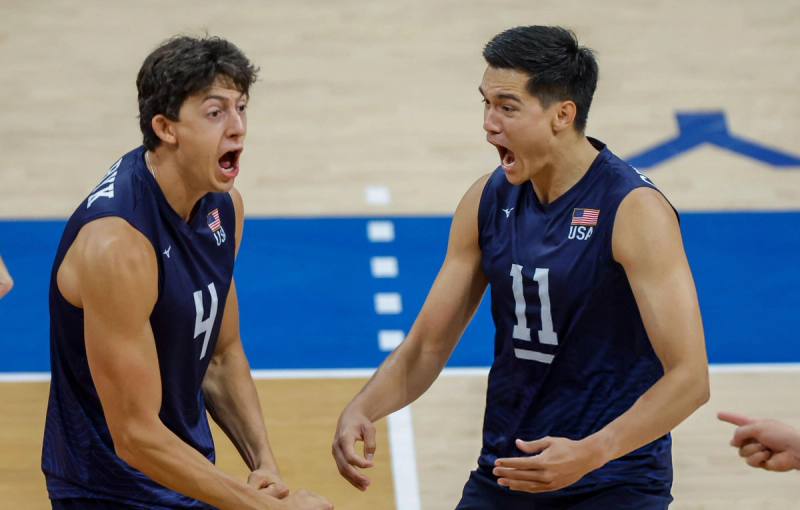 Two volleyball players in blue USA jerseys celebrate with expressions of excitement on a court.
