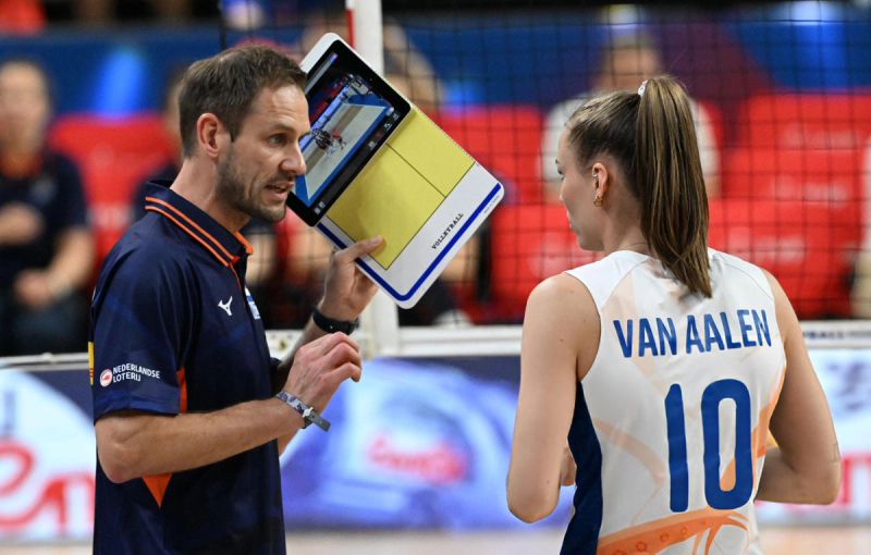 A volleyball coach is showing a tablet with a court diagram to a player during a game. The player is wearing a jersey with the name "Van Aalen" and the number 10.