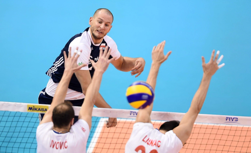 A male volleyball player in a dark jersey spikes the ball over the net as two opponents in white jerseys attempt to block it.