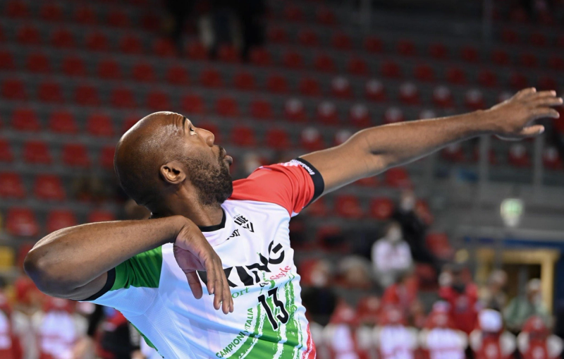A volleyball player in a multicolored jersey prepares to serve, with a blurred audience in the background.