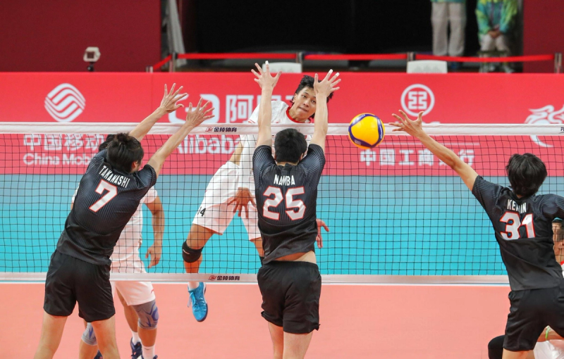 A volleyball player in a white uniform jumps to spike the ball over a net, while three opposing players in black jerseys attempt to block. The background features red advertising boards.