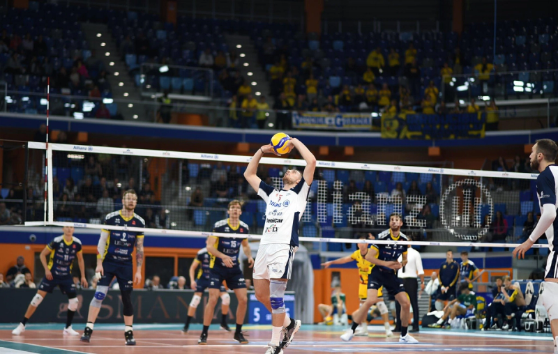 A volleyball player in a white uniform prepares to set the ball during a match. Players in yellow and blue uniforms are on the other side of the net. The court is surrounded by spectators in a large indoor arena.