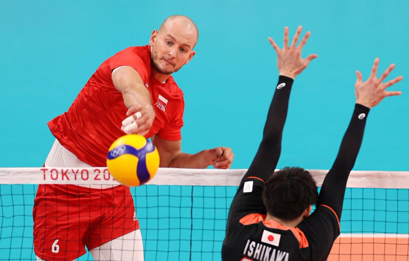 Two volleyball players compete at the net during a match. One player in a red jersey is about to hit the ball, while the other, in a black jersey, attempts to block. The net features "Tokyo 2020" text.