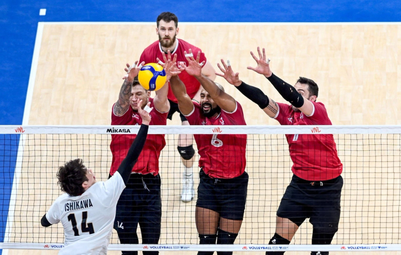 Volleyball match action showing a player in a white jersey attempting to spike the ball, while four players in red jerseys block at the net. The court is visible in the background.