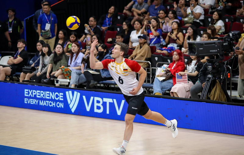 A volleyball player in a red and white jersey, with the number 6, prepares to serve during a match. The audience watches intently from the stands, and a camera is set up nearby.