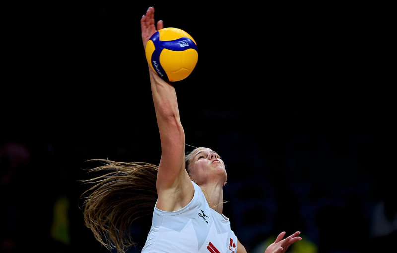 A volleyball player in a white jersey jumps high to spike a ball during a game, with a dark background.