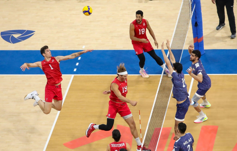 Volleyball match action shot. A player in a red uniform jumps to spike the ball over the net, while players in blue uniforms attempt to block. The court has a blue and wood pattern.