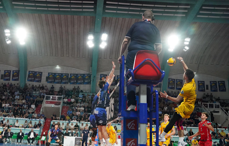 A volleyball player in a yellow jersey prepares to spike the ball over the net, while an opponent in a blue jersey attempts to block. A referee is seated on a chair next to the net. The background shows a packed audience in a sports arena.