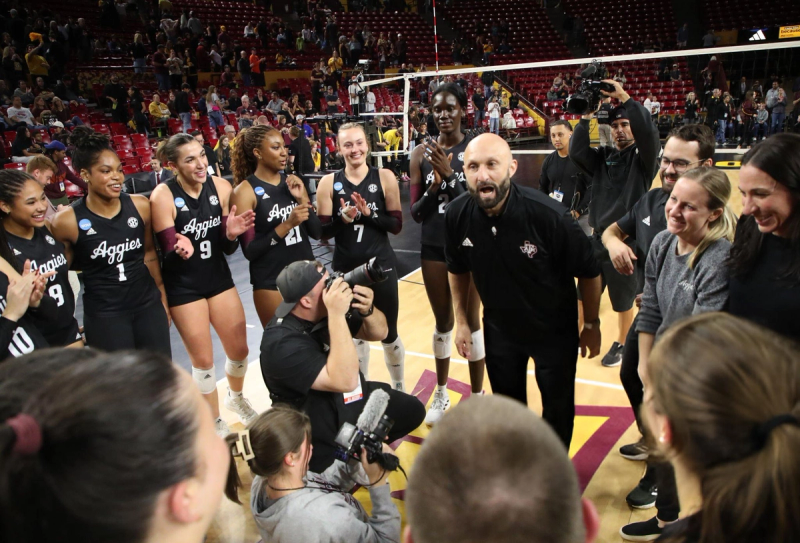 A group of volleyball players wearing black "Aggies" jerseys are gathered on a court, appearing to celebrate an event. A coach is speaking energetically to them, while photographers capture the moment. Spectators are visible in the background.