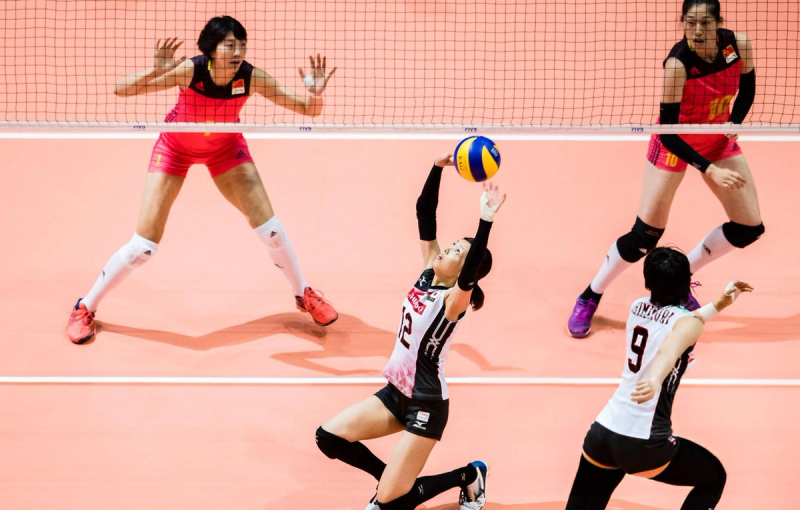 A volleyball player in a black and white uniform sets the ball, while two players in red-and-yellow uniforms prepare to block at the net. Another player in black and white is on the court ready to assist.