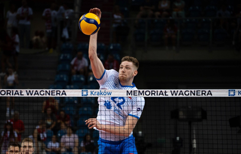 A volleyball player in a blue and white uniform is about to hit a ball over the net during a game, with "Memorial Wagner" visible on the net and spectators in the background.
