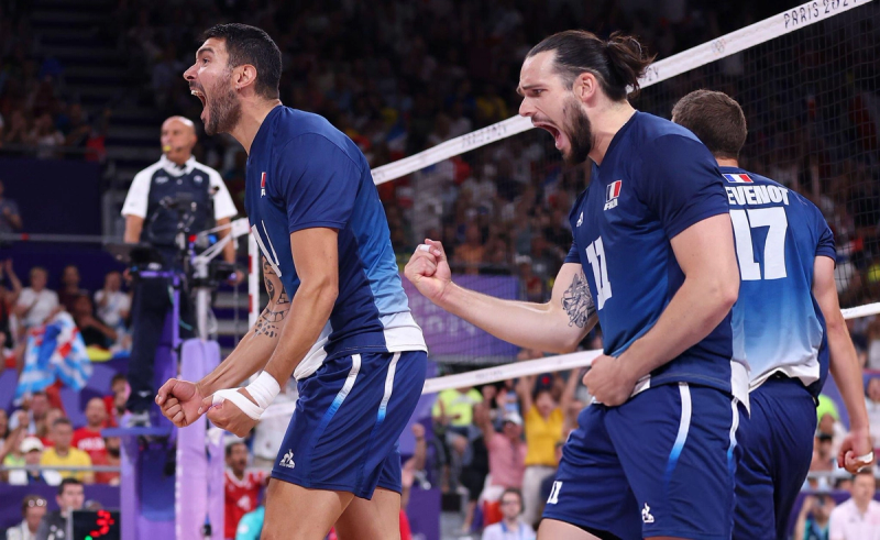 Two volleyball players in blue jerseys celebrate intensely on the court, fists clenched and mouths open in jubilation. A referee and spectators are visible in the background.