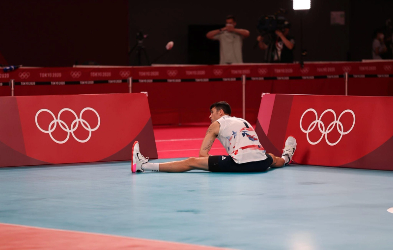 A volleyball player sits on the court with legs outstretched, leaning against a barrier with Olympic rings. The background shows a red spectator area and photographers.