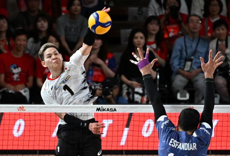 A volleyball player in a white jersey jumps to spike the ball against a blue-clad opponent at the net, with spectators in the background.