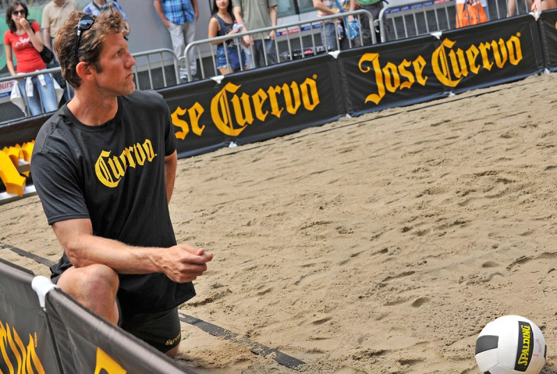 A man in a &quot;Cuervo&quot; t-shirt kneels on a sandy volleyball court with a Spalding ball nearby. Spectators stand behind a fence with &quot;Jose Cuervo&quot; banners.