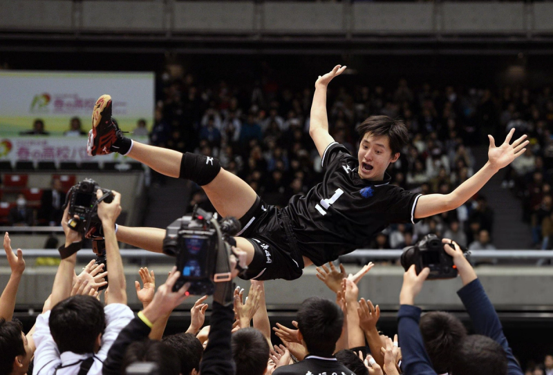 A volleyball player in a black uniform is joyfully tossed into the air by a crowd after a game, with photographers capturing the moment.