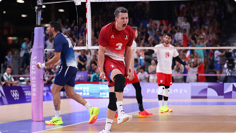 Volleyball player in red celebrates passionately on court, while teammates in background react. A player in blue walks past the net. Spectators fill the stands in the background.