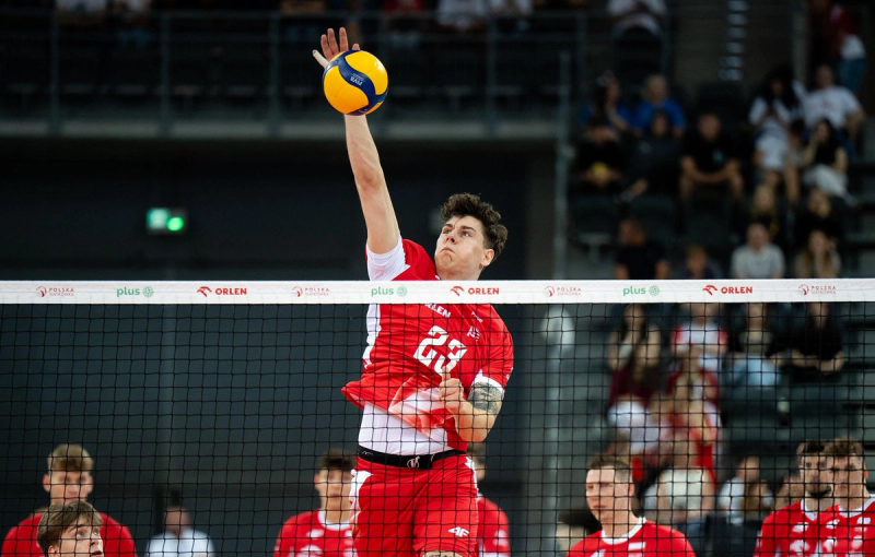 A volleyball player in a red jersey jumps to spike a ball during a match, with teammates and a crowd in the background.
