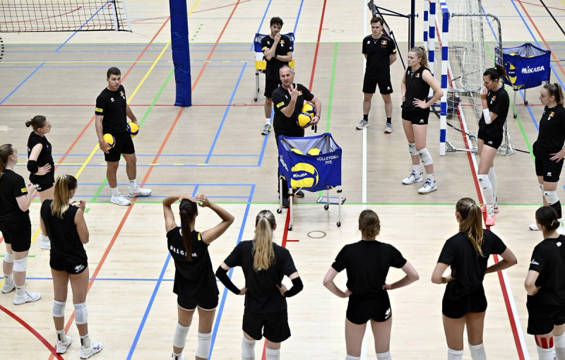 A volleyball coach instructs a team of players in black uniforms on an indoor court, with volleyballs in carts nearby.