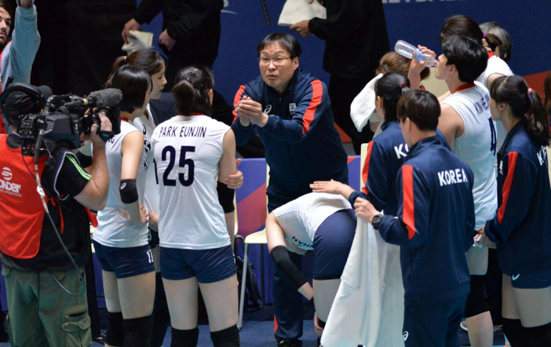 A volleyball coach gives instructions to the team during a huddle. Players are wearing white jerseys with their names and numbers, while a camera crew records the scene. Some players drink water or listen intently.