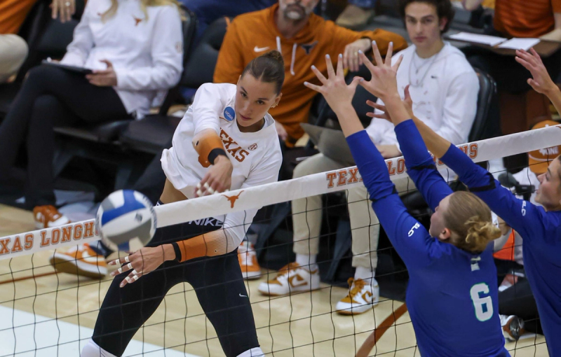 A volleyball player in a white jersey jumps to spike the ball over the net, while two players in blue attempt to block the shot. The scene takes place in an indoor court with spectators in the background.