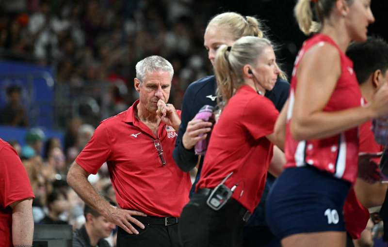 A group of people in a sports setting, wearing red and blue uniforms with "USA" on them. One person is deep in thought with their hand on their chin, while others are engaged in conversation or activity.