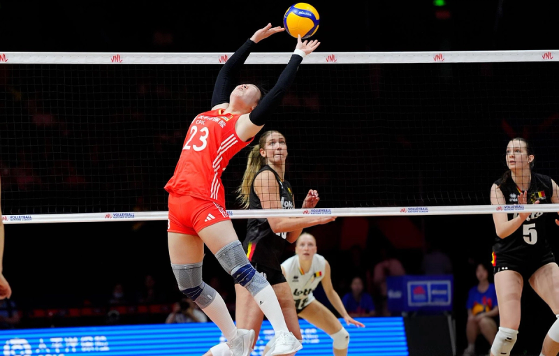 A volleyball player in red prepares to set the ball during a match, with opponents in black and additional players in the background. The setting is a professional indoor volleyball court.