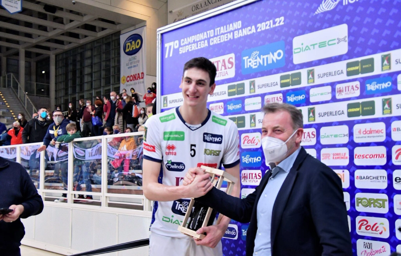 A volleyball player in a white jersey receives a trophy from a man in a suit in front of a sports event backdrop. Fans are visible in the background.