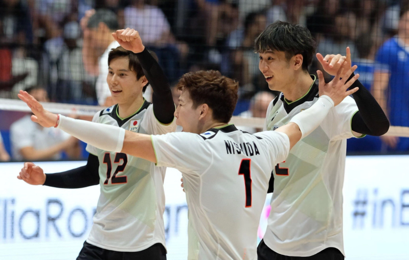 Three volleyball players in white jerseys, marked with Japanese flags, celebrate on the court with smiles and raised arms.