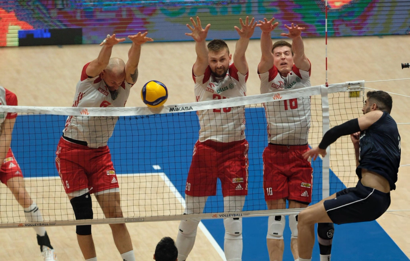 Three volleyball players in red uniforms jump to block the ball at the net, while a player in a dark uniform prepares to spike.