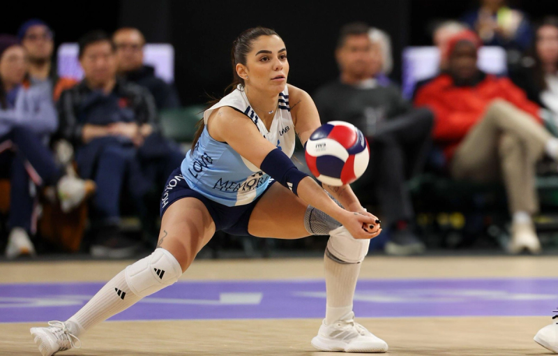 A volleyball player in a blue and white uniform crouches low to pass a volleyball during a game, with spectators seated in the background.