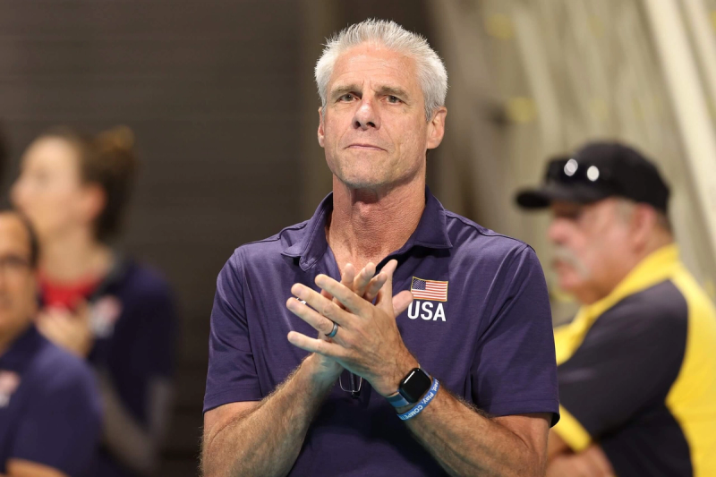 A man with short gray hair is clapping while wearing a navy shirt with a USA flag. The background shows other people slightly out of focus.