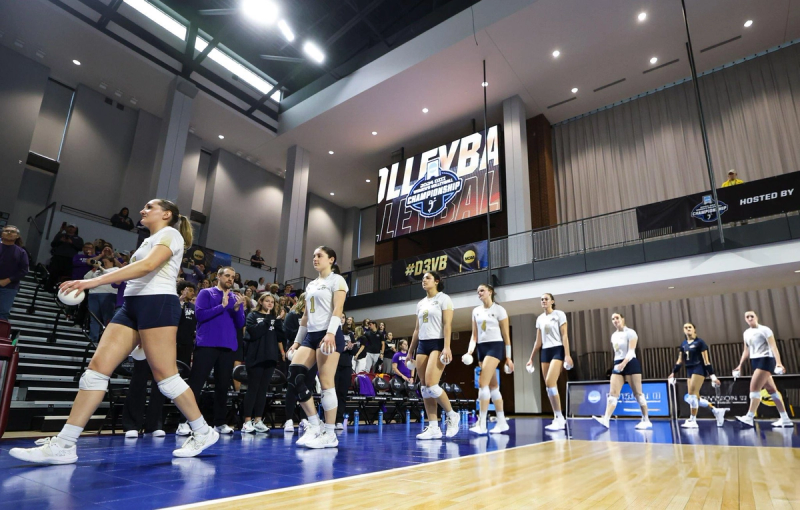 A women's volleyball team enters an indoor stadium, ready for a match. They're wearing white jerseys and navy shorts. A large banner reading "Championship" hangs overhead, and spectators are seen in the background clapping.