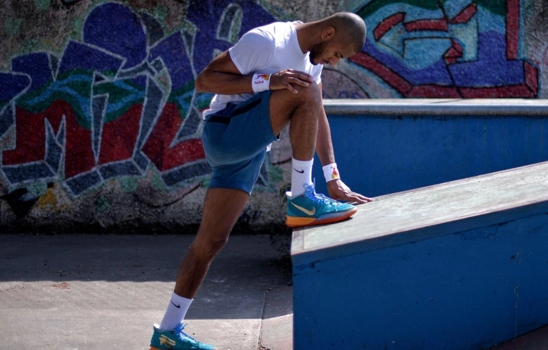 A man in athletic attire is stretching his leg on a ramp in an urban environment with colorful graffiti in the background.