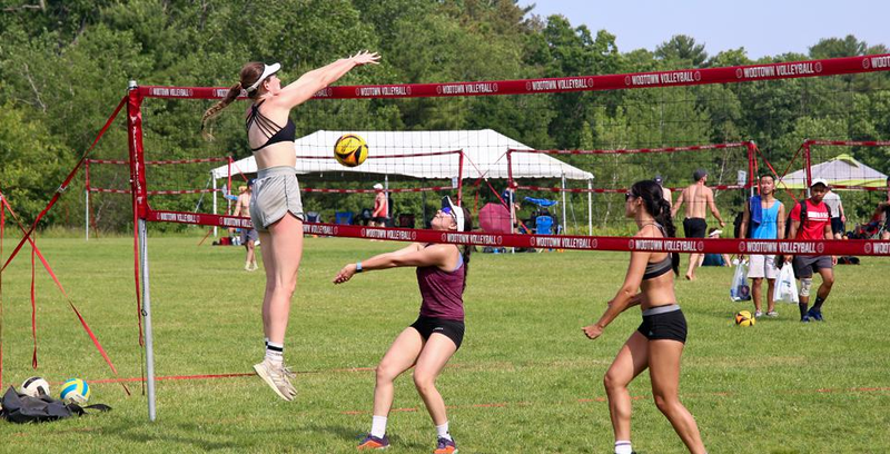 Three women playing beach volleyball on grass, with one jumping to hit the ball. Spectators and other players are in the background.