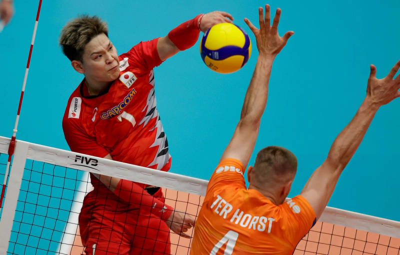 A volleyball player in a red jersey is spiking the ball over the net, while an opposing player in an orange jersey attempts to block.