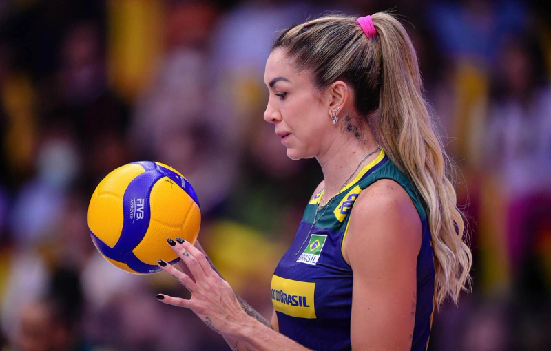 A volleyball player in a Brazil uniform with a ponytail holds a volleyball, focusing intently before serving.