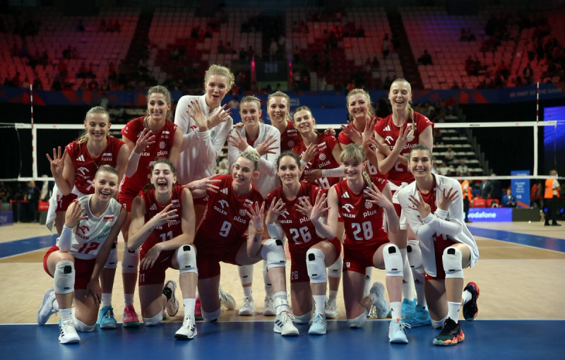 A women's volleyball team posing on the court. Many are wearing red uniforms, and they are smiling and gesturing with their hands as they kneel and stand together. The volleyball net and an audience are visible in the background.