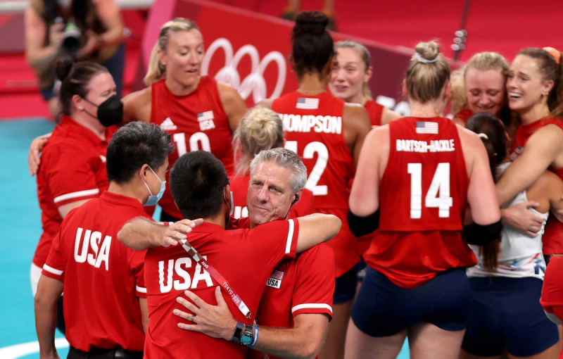 A group of athletes and coaches in red "USA" uniforms share an emotional hug on a volleyball court with the Olympic rings in the background.