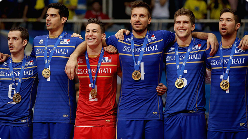 A group of six volleyball players wearing medals and team uniforms stand arm-in-arm, smiling. The uniforms are blue, except for one player in red.