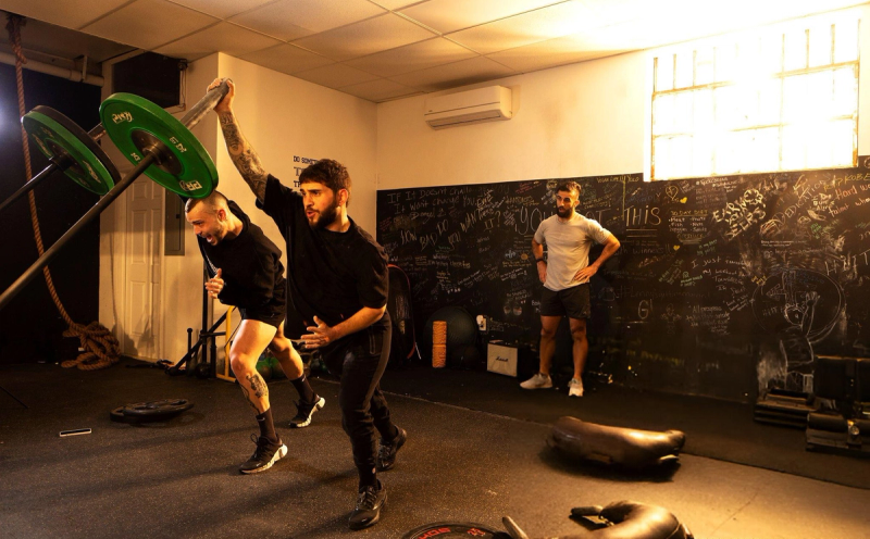 Two people are engaged in a dynamic exercise with a barbell equipped with green weights in a gym. One person is actively pushing the barbell, while the other supports him. A third person stands in the background, observing with hands on hips. The walls are decorated with motivational quotes and writings.