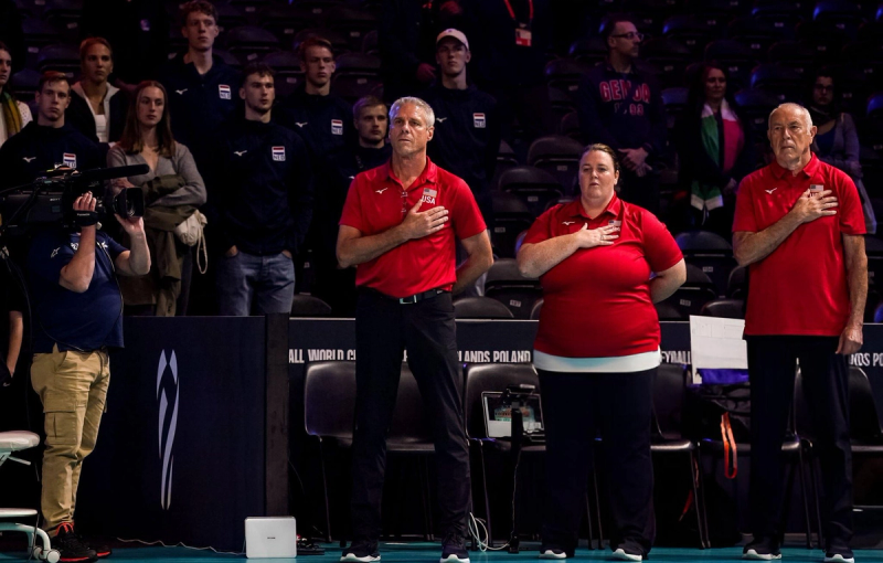 Three people wearing red shirts stand with hands over their hearts during a ceremony. A camera operator films them, and a group of people watch from the background.
